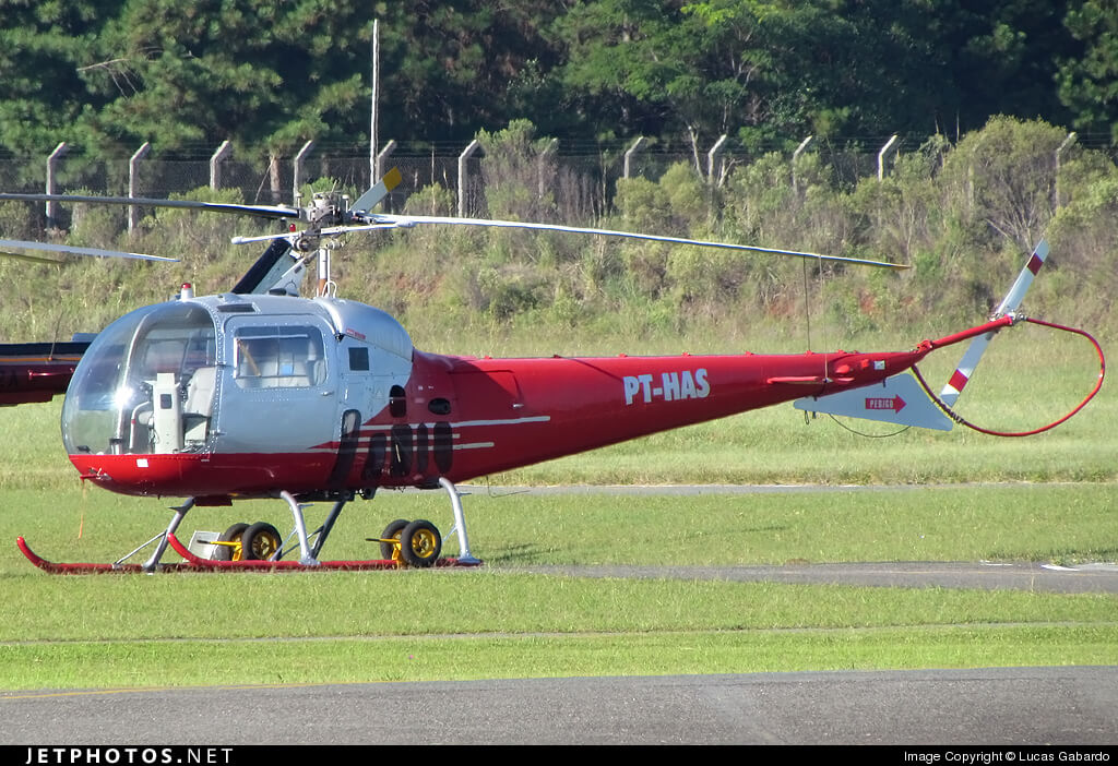 Um helicóptero vermelho e prateado com a matrícula "PT-HAS" está estacionado em uma pista de grama, com uma cerca e árvores ao fundo.