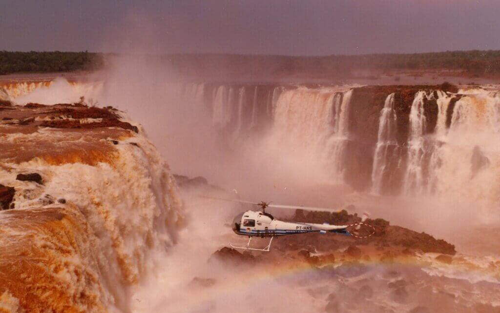 Um helicóptero branco e azul pousado em uma área rochosa em frente a uma grande cachoeira, com um arco-íris visível na névoa abaixo.