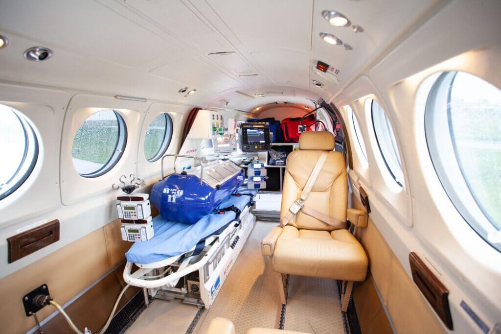 Uma maca com lençóis azuis e equipamentos médicos em um avião, com uma poltrona bege ao lado.