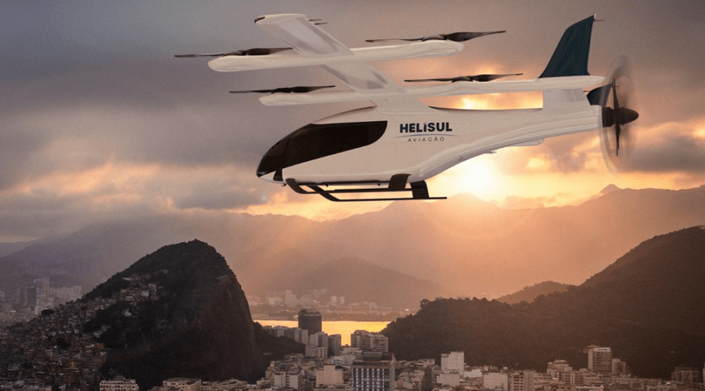 Uma aeronave branca com a inscrição "HELISUL AVIAÇÃO" está voando sobre uma cidade com montanhas e um pôr do sol alaranjado ao fundo. A aeronave possui múltiplas hélices e um design moderno.