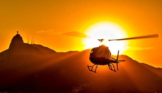 Um helicóptero em silhueta voa contra um sol poente ou nascente laranja e amarelo, com o Cristo Redentor e montanhas do Rio de Janeiro ao fundo.