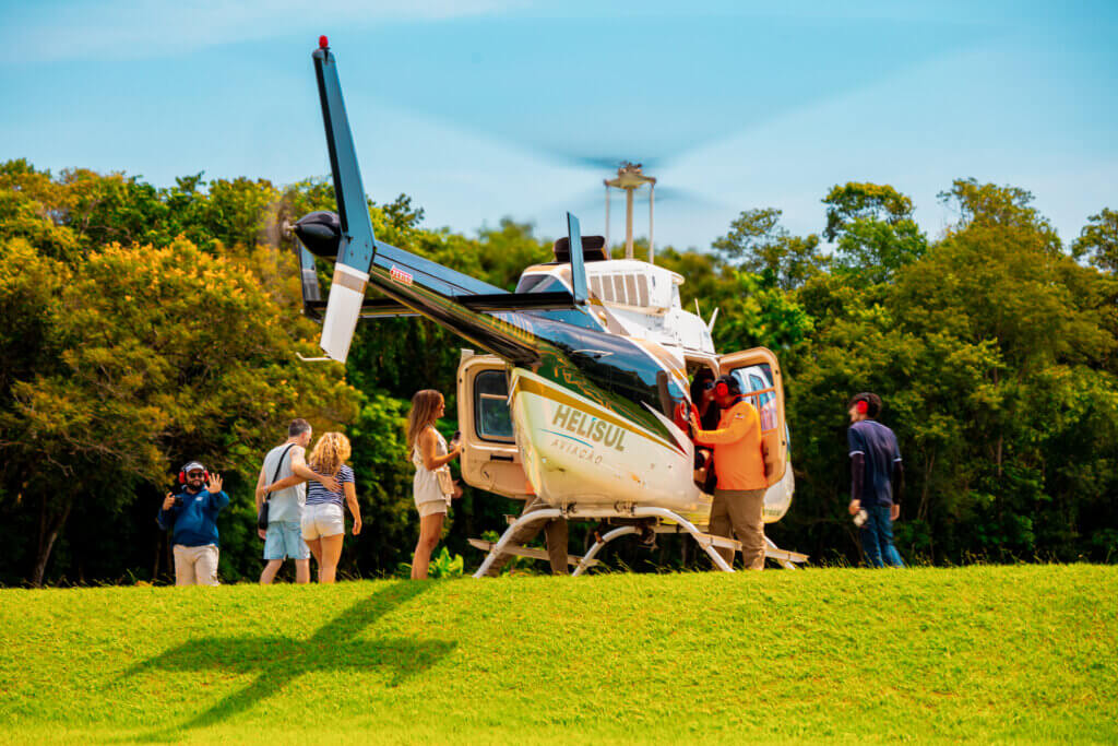 Helicóptero da helisul aviação pousado em um gramado verde com passageiros e equipe técnica ao redor sob um céu azul claro.