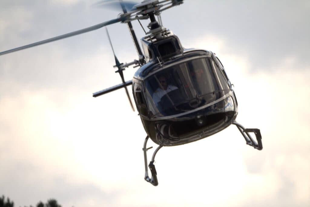 Visão frontal de um helicóptero de cor escura em pleno voo, com piloto e copiloto visíveis na cabine contra um céu nublado.